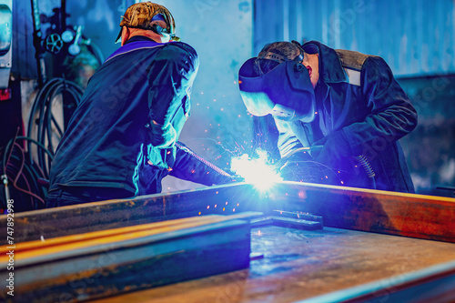 A welder wearing a protective mask for metal welding and protective gloves performs welding work at a metal structures plant. Welding a steel industrial beam. Sparks, hot flame from welding.