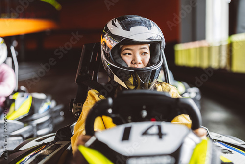 young asian female kart racer with helmet holding the steering wheel in her racing car on the recreational circuit © Aida