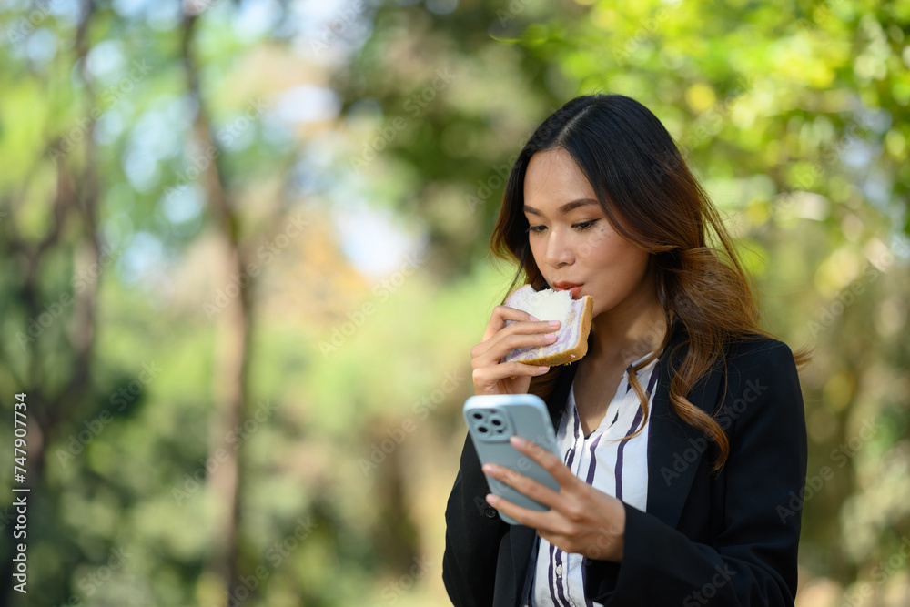 Businesswoman having lunch break eating sandwich and using mobile phone in the park Businesswoman having lunch break eating sandwich and using mobile phone in the park