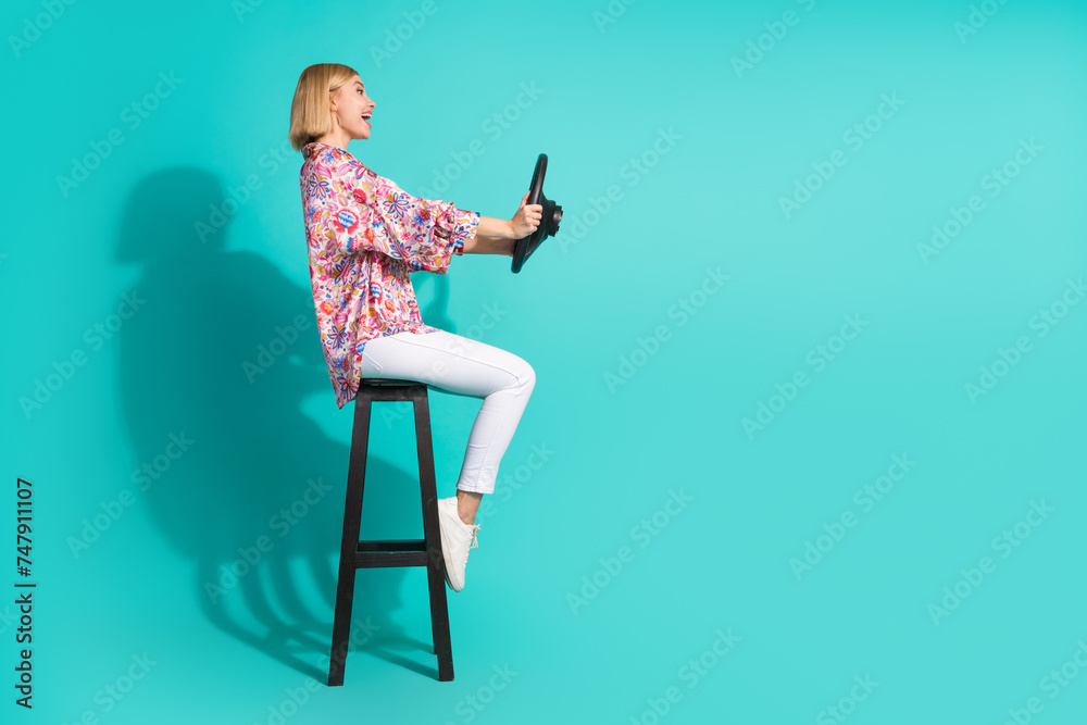 Full body photo of pretty young girl sit chair hold steering wheel wear trendy pink print outfit isolated on cyan color background Full body photo of pretty young girl sit chair hold steering wheel wear trendy pink print outfit isolated on cyan color background