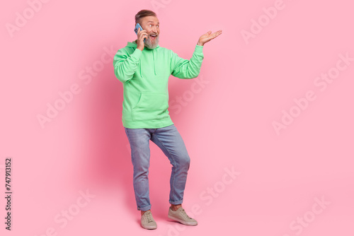 Full length photo of handsome senior man talking mobile phone roaming dressed stylish green garment isolated on pink color background