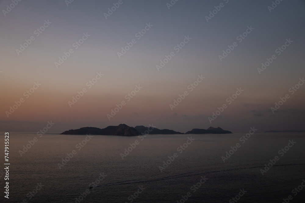 Calm seas at dusk: shot from Monteferro capturing the Atlantic Ocean and Cies Islands' silhouette in the center. Low gray clouds contrast with the orange sky, as a lone boat cruises into twilight.