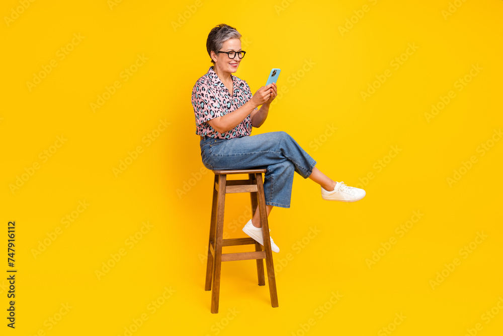 Full body photo of attractive pensioner woman hold gadget sit chair wear trendy leopard print clothes isolated on yellow color background