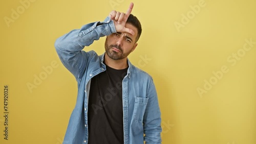 Cheeky young hispanic man in denim shirt, standing against yellow background, insulting people with loser gesture - fingers to forehead, mocking and laughing rude tease