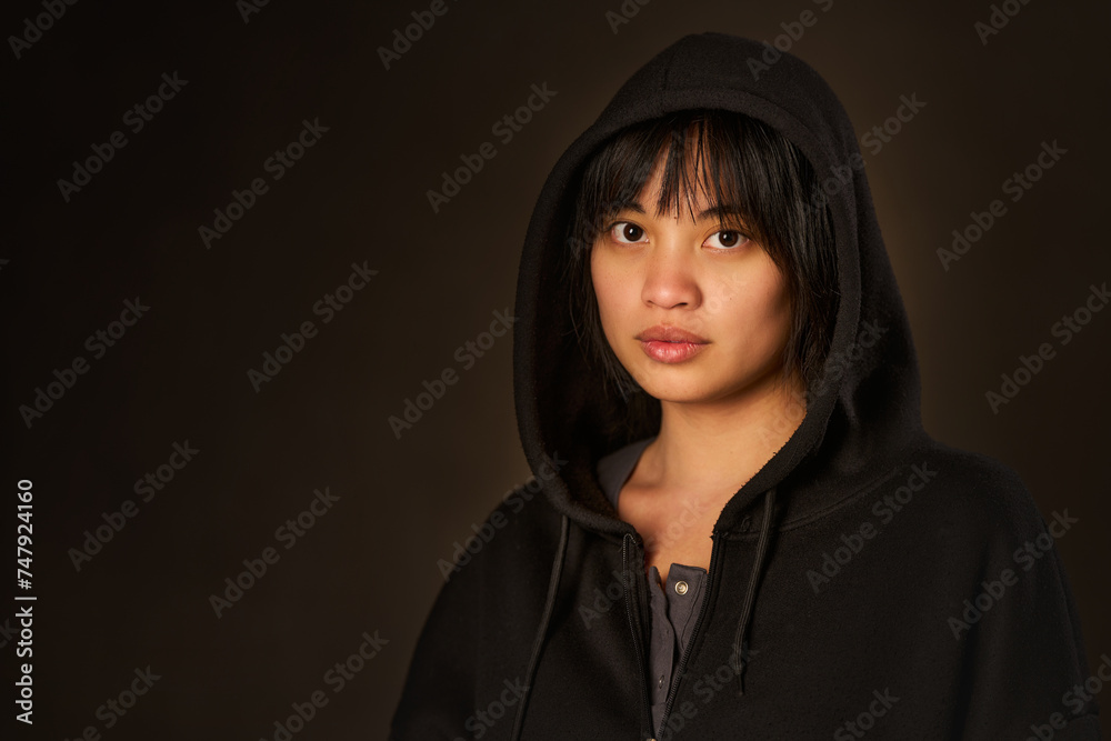 Fototapeta premium Mysterious portrait of a young Asian woman in a black hoodie and sweatshirt with a dark background