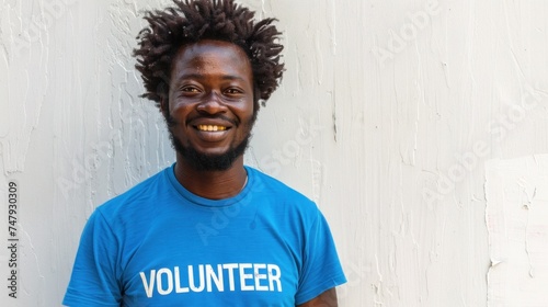 Smiling man with curly hair wearing a blue t-shirt with the word "VOLUNTEER" in white letters standing against a white wall.