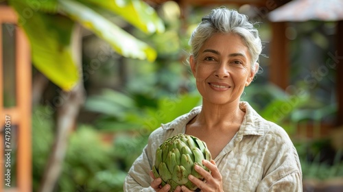 A woman with gray hair smiling holding a large green artichoke surrounded by lush greenery.
