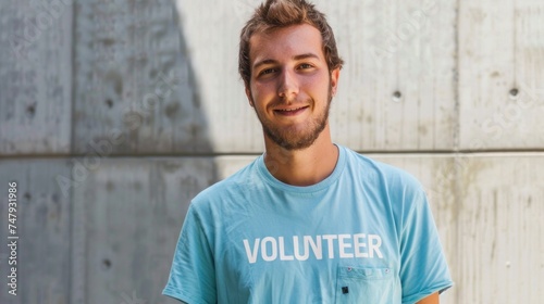 A young man with a beard wearing a light blue t-shirt with the word "VOLUNTEER" printed on it smiling at the camera against a textured concrete wall.