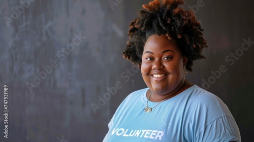 Smiling woman with curly hair wearing a blue t-shirt with the word "VOLUNTEER" on it standing against a textured gray background.