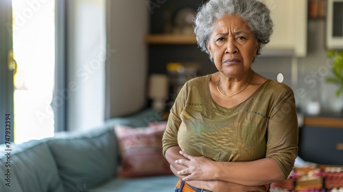 An elderly woman with gray hair wearing a green top standing in a living room with a concerned expression her hands on her stomach.