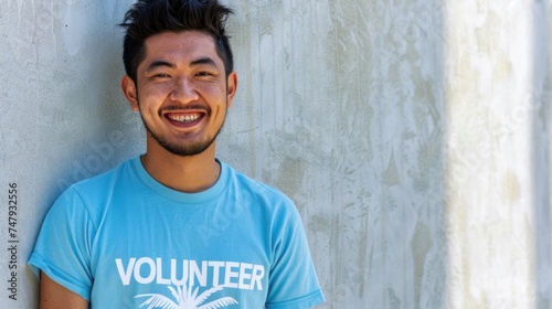 A young man with a beard and short hair wearing a blue t-shirt with the word "VOLUNTEER" printed on it smiling against a textured concrete wall.