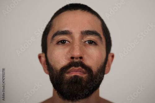 A studio portrait of a multiracial Jewish, Asian, and White man looking neutrally at the camera. His blank expression and shirtless pose highlight his strong eyebrows, dark beard, and dark hair. © Levi Meir Clancy