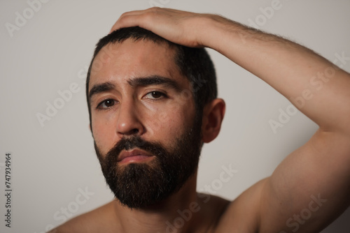 With his brow slightly furrowed, a man lifts his arm up and puts his arm on his head. He is shirtless, and his upper arm is visible, expressing a mix of seriousness, vulnerability, and intimacy.