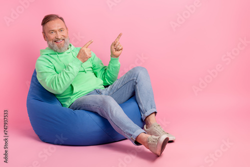 Full size photo of cool pensioner man sit bean bag point empty space dressed stylish green clothes isolated on pink color background