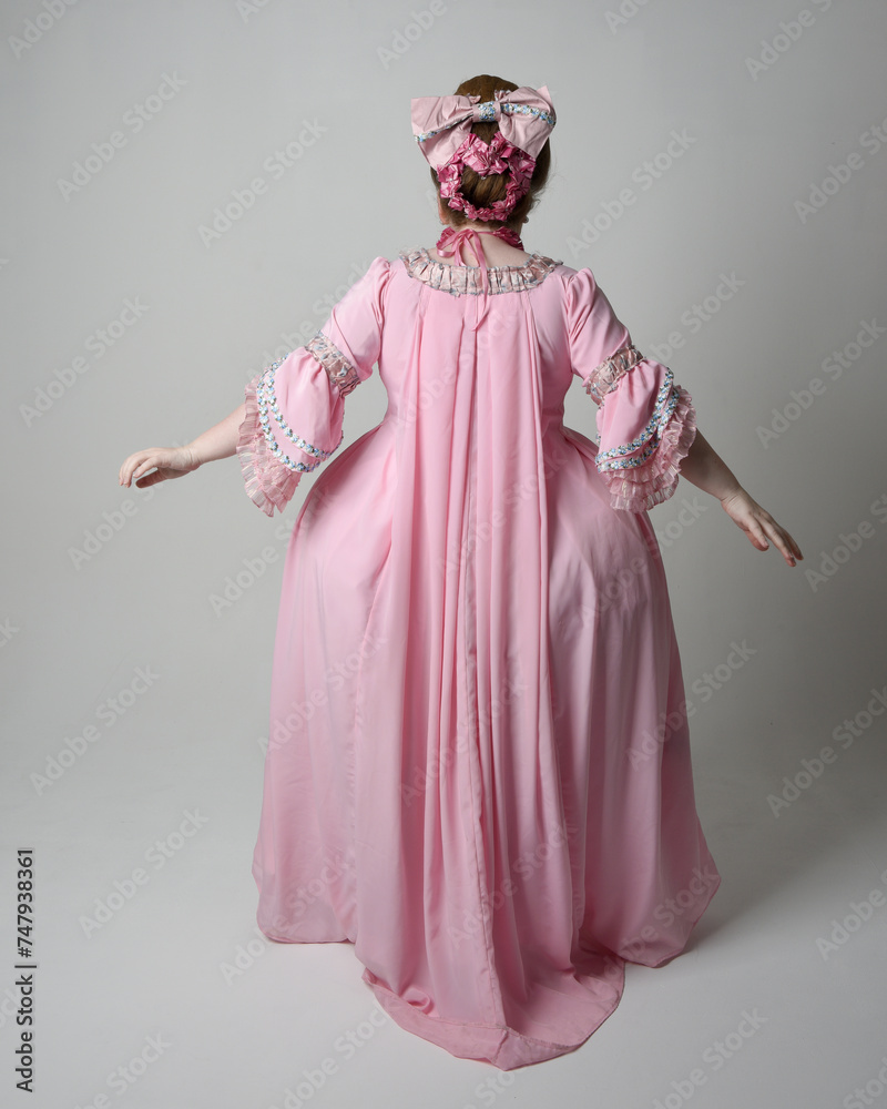 Full length portrait of woman wearing historical French baroque pink gown in style of Marie Antoinette with elegant hairstyle. Standing pose, walking away from the camera, isolated on studio backgroun
