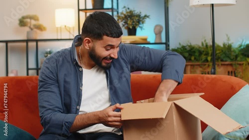 Excited young Indian man unpacking delivery parcel sitting on sofa in living room. Smiling Hispanic guy shopper online shop customer clenching fists receiving purchase by fast postal shipping courier. photo