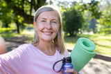 Healthy mature woman with a yoga mat and water bottle, smiling in a sunlit park. Fitness and wellbeing concept.