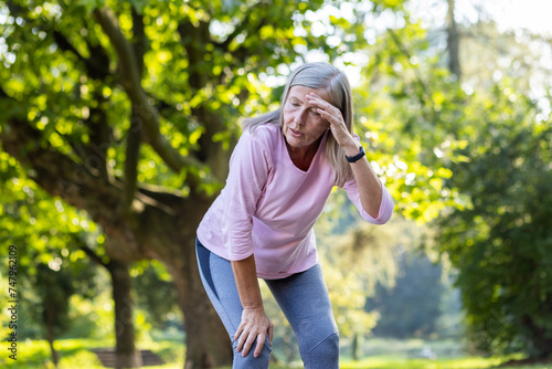 Quadro su tela An active senior woman taking a break from running, appears tired, wiping sweat from her forehead in a peaceful park