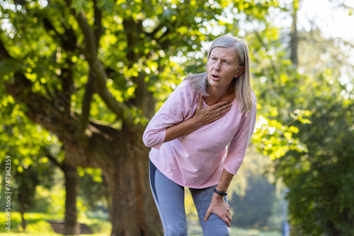Fotografie An elderly woman pauses her workout in the park, looking distressed with a hand over her chest, possibly indicating chest pain