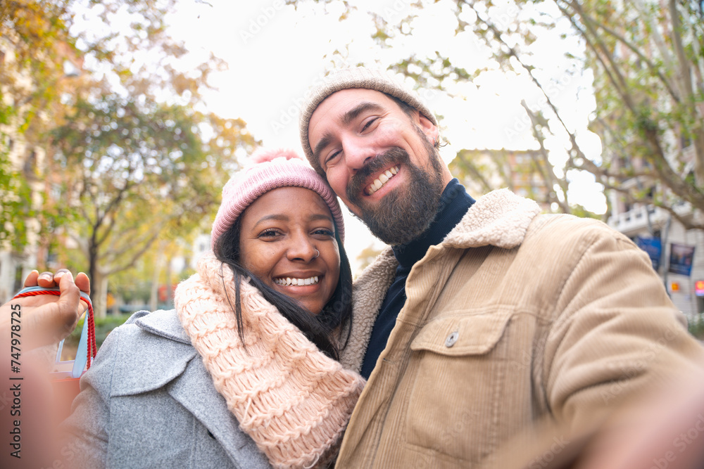 Fototapeta premium Multiethnic smiling young couple with shopping bags making selfie on cell phone outdoors.