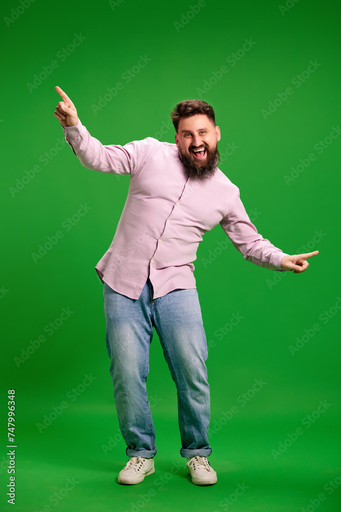 Full length portrait of young man dancing with delightful expression against vibrant green studio background. Time of success. Concept of human emotions, self-expression, rest, hobbies, leisure.