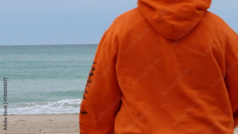 Mujer joven con sudadera naranja y estilo alternativo caminando hacia la orilla en la Playa de San Juan de Alicante, España