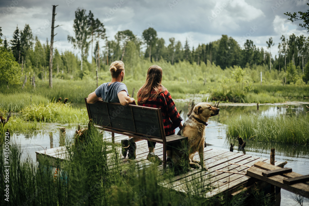 couple with dog sitting on bench in front of stomp lake surrounded by wild forest couple with dog sitting on bench in front of stomp lake surrounded by wild forest