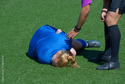Injured girl football player lying on the soccer field double up with pain, referee puts hand to calm her