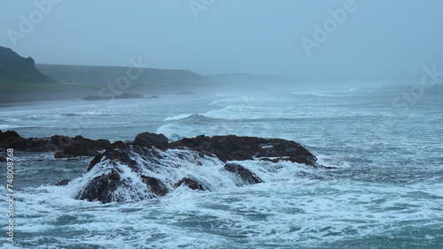 Beautiful waves break on rocks of rocky shore. Clip. Stone shore with storm waves in cloudy weather. Dramatic landscape of stone coast with beautiful splashes of waves
