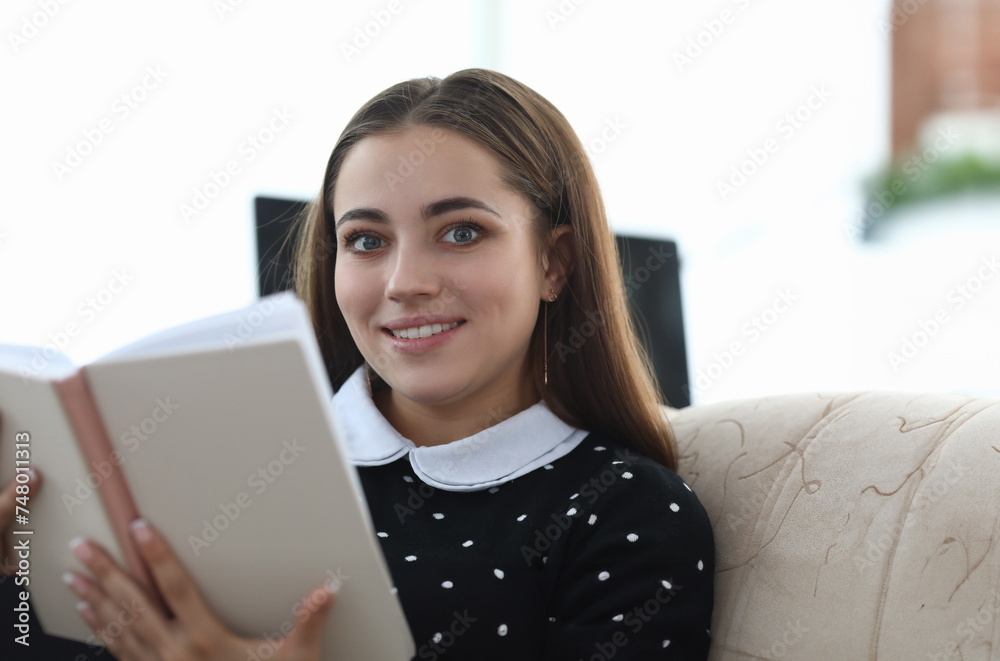 Portrait of beautiful young woman in cozy home interior sitting on sofa and reading personal diary. Charming smiling brunette holding notebook. Leisure concept Portrait of beautiful young woman in cozy home interior sitting on sofa and reading personal diary. Charming smiling brunette holding notebook. Leisure concept