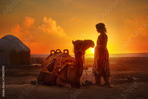 Silhouette of woman in traditional national clothes looks at beautiful sunset in desert next to yurt and camel. © soft_light