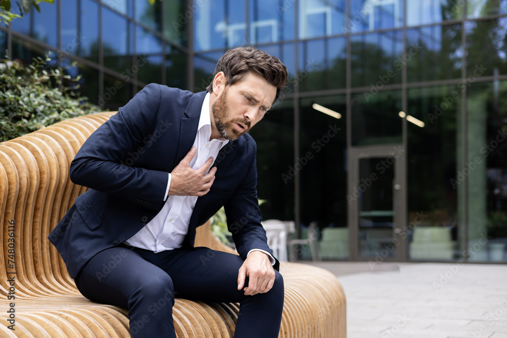 A young businessman man is sitting on a bench near the office and holding to his chest, having a heart attack, feeling severe pain and suffering from difficulty breathing, he needs medical help A young businessman man is sitting on a bench near the office and holding to his chest, having a heart attack, feeling severe pain and suffering from difficulty breathing, he needs medical help