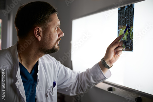 Side view of serious doctor studying MRI results. Male specialist working in hospital, carrying out description of radiographs of patient, pointing by pen. Concept of medicine and hospital.