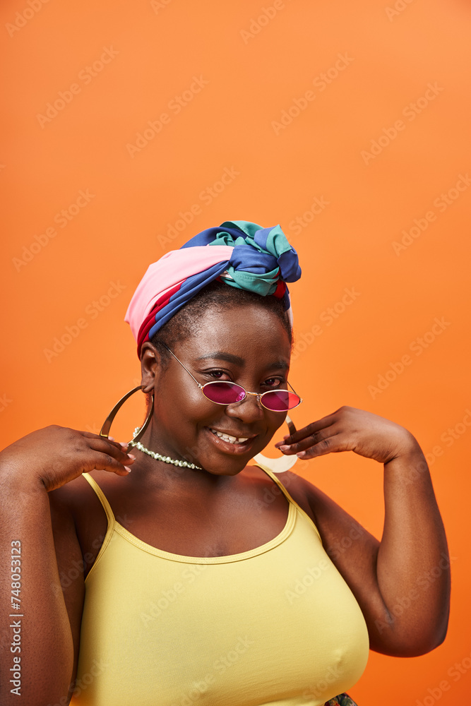 Fashionable plus size and happy african american woman in headscarf and trendy sunglasses on orange Fashionable plus size and happy african american woman in headscarf and trendy sunglasses on orange