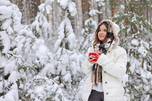 Młoda ładna dziewczyna trzyma w dłoniach czerwony kubek z gorącą herbatą. Spaceruje w mroźny zimowy dzień po lesie.
