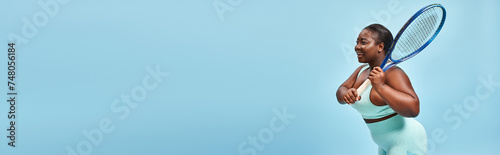 plus size dark skinned woman holding tennis racket on blue backdrop, body positive and sport banner © LIGHTFIELD STUDIOS