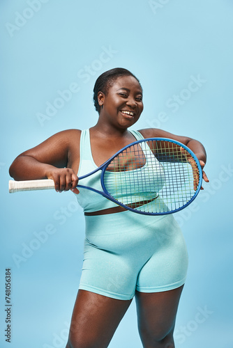 happy plus size dark skinned woman holding tennis racket on blue backdrop, body positive and sport © LIGHTFIELD STUDIOS