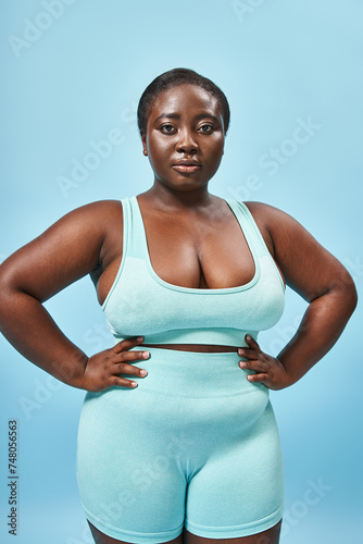 strong plus size african american woman in blue sportswear with hands on hips on matching backdrop