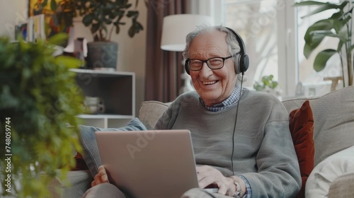 A happy elderly man with a headset uses his laptop at home, sitting on his couch, looking at the computer screen, watching a video lesson, and copying the text.