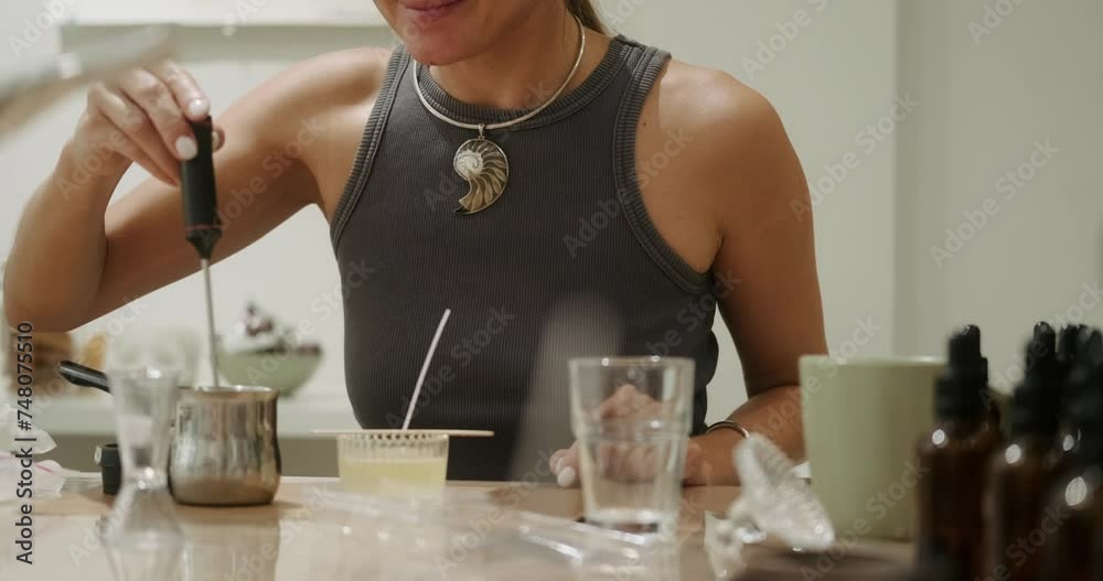 A young woman during a class on making scented candles in a workshop. A woman stirs liquid wax in a jar using a thermometer and smells the resulting aroma. The process of making a handmade candle.