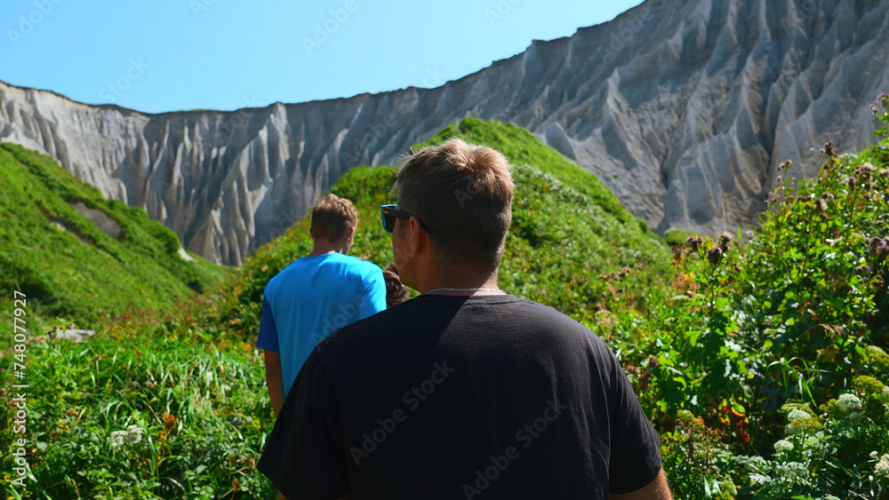 Group of people walks along hiking trail with rocks in summer. Clip. Beautiful tourist route with path and green grasses to white rocks. Amazing trail with tourists at white rocks on sunny summer day