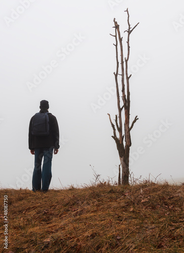 Young man in jacket and trousers with bacpack standing on hill with dead tree in morning fog. Czech landscape, travel, relax concept © Space Creator