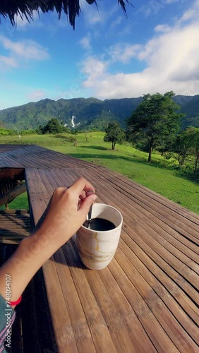 A hand stirring a cup of coffee on a bamboo balcony with a lush countryside summer landscape and mountains in the background showing the candid daily life in rural Philippines