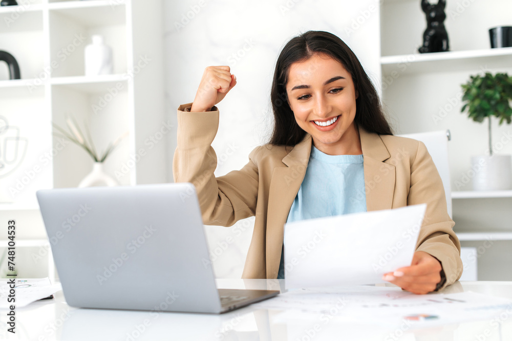 Success, profit. Happy indian or arabian business woman, ceo, financial manager, secretary, sit in modern office with laptop, studying documents, rejoicing in a good results, gesturing hand, smiling Success, profit. Happy indian or arabian business woman, ceo, financial manager, secretary, sit in modern office with laptop, studying documents, rejoicing in a good results, gesturing hand, smiling