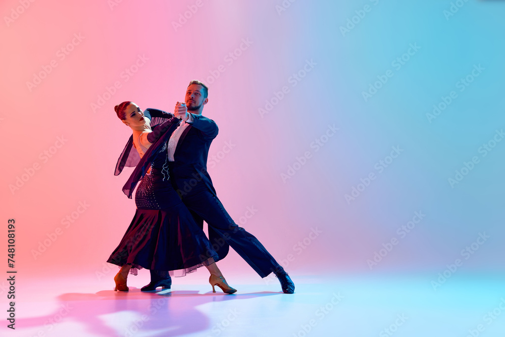 Fototapeta premium Young man and woman, talented ballroom dancers in motion, dancing in black costumes against gradient pink blue background in neon light. Concept of dance class, hobby, art, dance school, talent