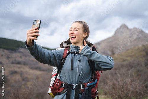 Smiling laughing hiker woman with backpack showing thumb up gesture while making selfie, camper with tourist gears expressing positivity in the mountains, journey tourism adventure