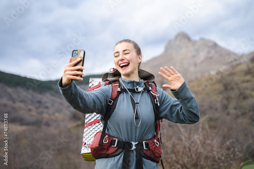 Smiling laughing hiker woman taking selfie, traveller speak to camera wave hand against the backdrop of the rocky mountains, hiking and climbing cliff, journey tourism adventure sport concept photo