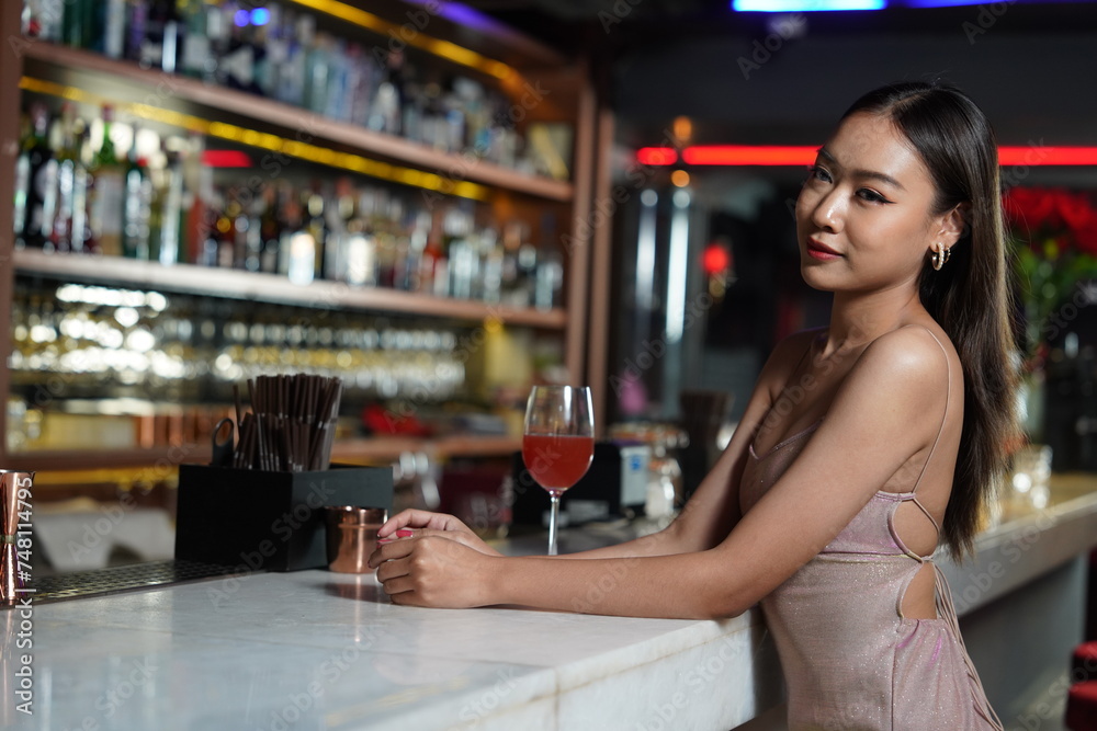 Beautiful Asian woman wearing a white dress holding a glass of red wine drinking a cocktail at the bar counter, partying in a nightclub
