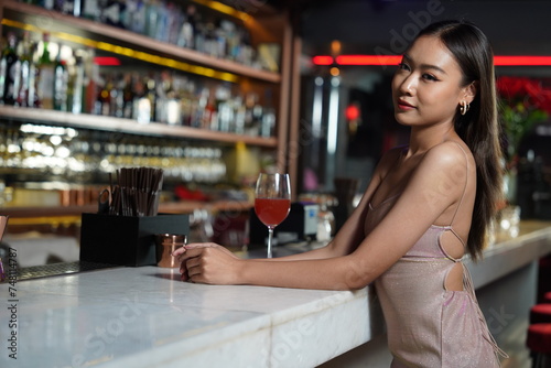Beautiful Asian woman wearing a white dress holding a glass of red wine drinking a cocktail at the bar counter, partying in a nightclub
