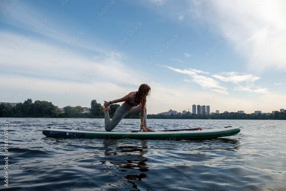 Young woman balances on SUP board stretching legs to engage muscle groups. Lady relaxes gliding on river surface on paddleboard
